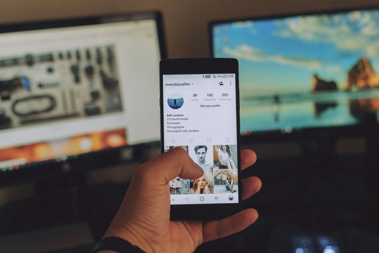 Holding cell phone, browsing Instagram, in front of two computer screens.