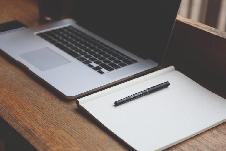 Wooden desk with a laptop sitting on top with a open notepad and pen beside it.