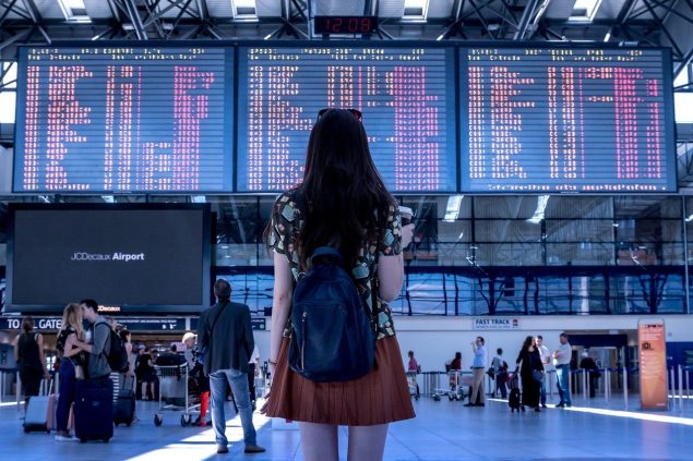 Woman with backpack in an airport facing a billboard with departures and arrivals.