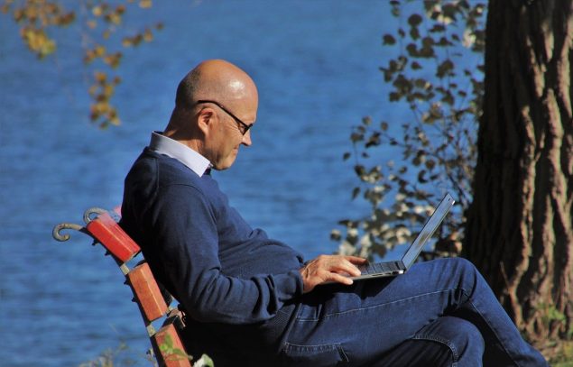 Elderly man sitting on a bench, using a laptop computer next to the water.